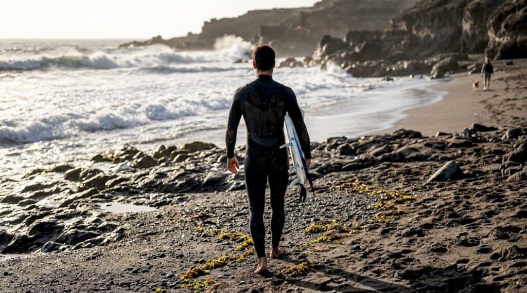 Un surfista se prepara para adentrarse en el mar desde una playa de arena negra volcánica.